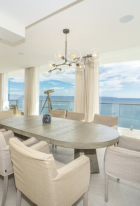 A dining area with an oval stone table and beige chairs, featuring a sculptural chandelier and a large glass-walled balcony.