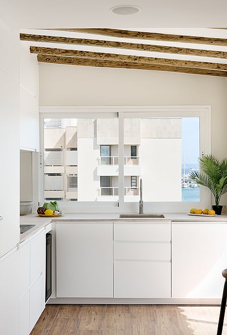 Modern kitchen with white cabinets, wood ceiling beams, and a window overlooking an urban exterior.