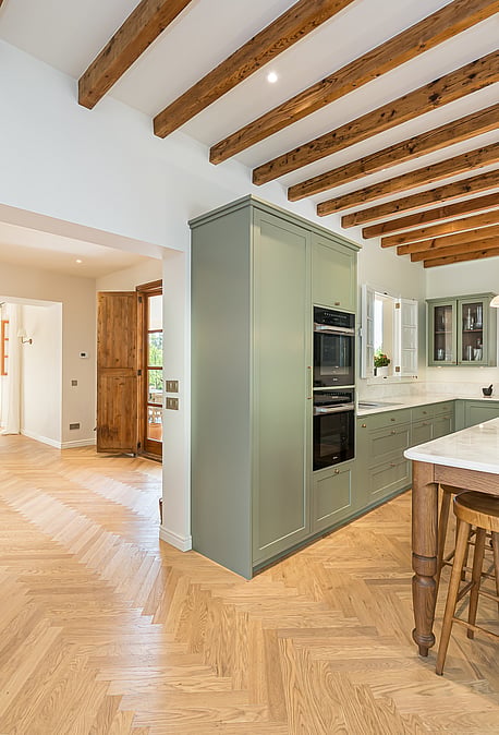 Modern kitchen featuring sage green cabinets, herringbone wood floors, a marble-top island, and exposed timber ceiling beams.