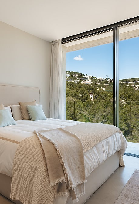 Modern bedroom with floor-to-ceiling windows overlooking green hills, featuring neutral textiles and a wooden side table.