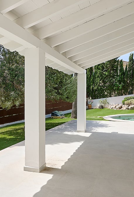 Terraza cubierta blanca con vigas de madera frente a césped artificial, piscina circular, pinos y cipreses