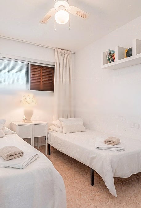 A bright guest bedroom featuring two single beds, light-toned textiles, a white wall-mounted shelf, and a minimalist aesthetic.