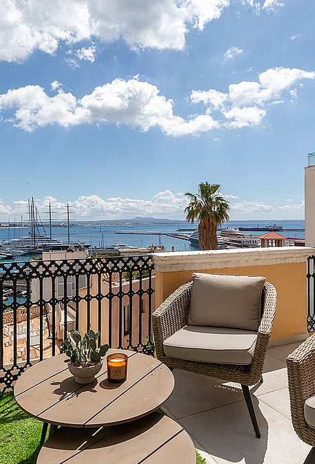 Outdoor terrace with woven armchairs, a round wooden table, and iron railings overlooking a marina and sea under blue sky.