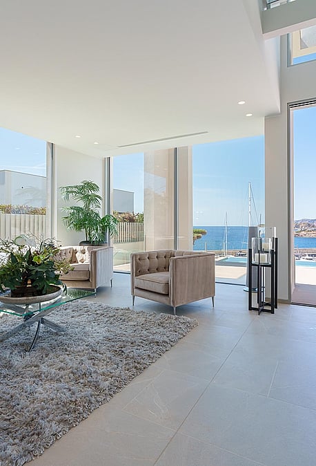 Bright living room with white walls, tufted beige sofas, glass coffee table, and large windows looking out to the harbor.