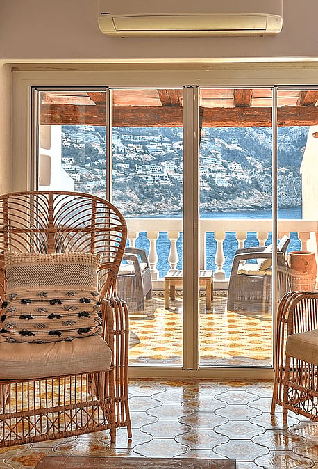 Two rattan armchairs facing sliding glass doors that open onto a sea-view terrace, with decorative mirrors and wall clock.