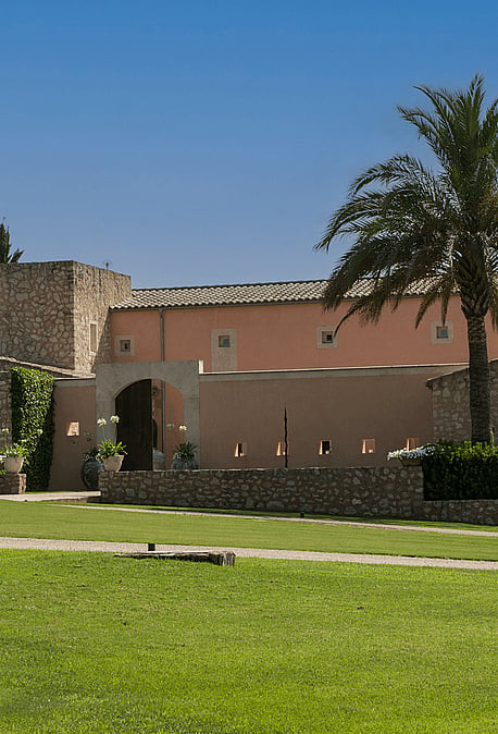 Exterior view of a traditional stone and peach-colored finca with arched entrance, large palm tree, and manicured green lawn.