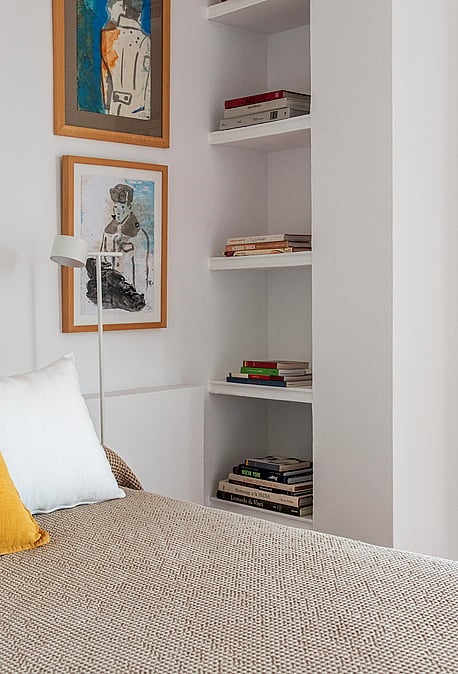 A close-up of a bedroom featuring a textured beige bedspread, a bright yellow throw pillow, and integrated white wall shelving.