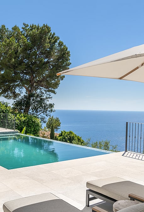 A rectangular swimming pool on a pale stone terrace overlooks the blue sea, framed by a pine tree and lounge furniture.