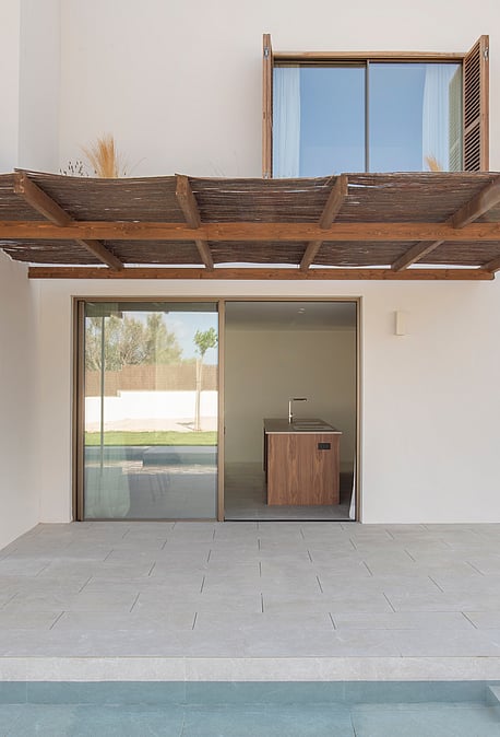 A stone terrace with a wooden pergola sits beside a swimming pool, leading to a modern white villa with glass doors.