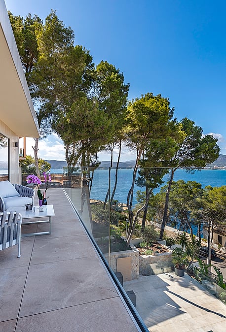 A wide terrace with grey outdoor furniture, glass railings, and a view overlooking the Mediterranean coastline and pines.