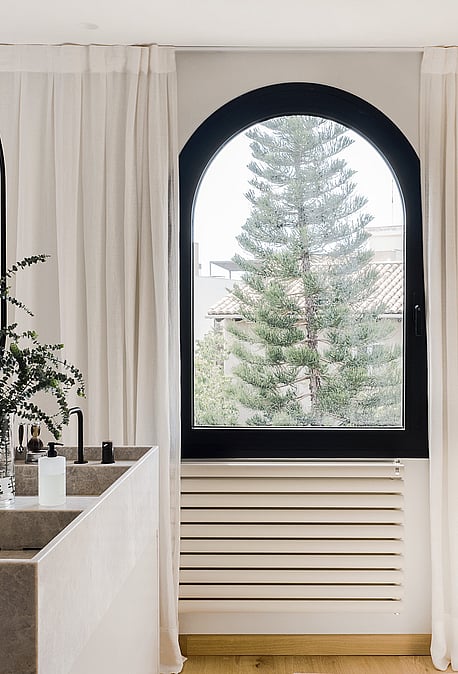 Zwei Waschbecken aus Stein mit schwarzen Armaturen, zwei ovale Hängespiegel reflektieren das Bogenfenster und Pinienausblick