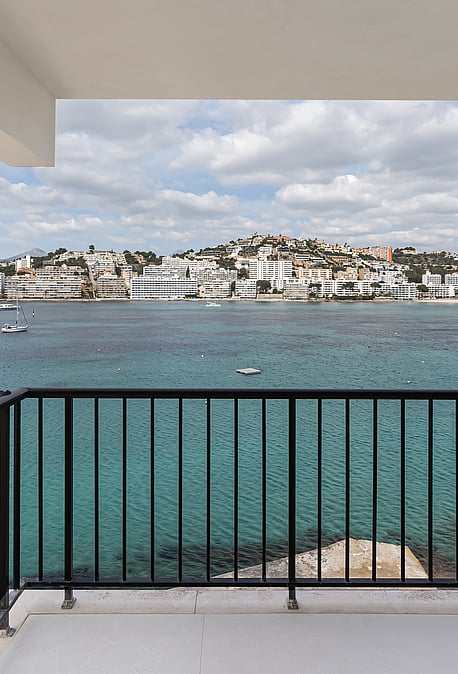 A balcony with black metal railing overlooking a turquoise bay and white coastal town under a soft sky.