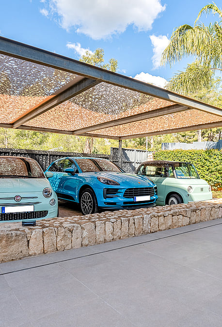 Covered carport with steel frame and patterned shade canopy beside a natural-stone wall.