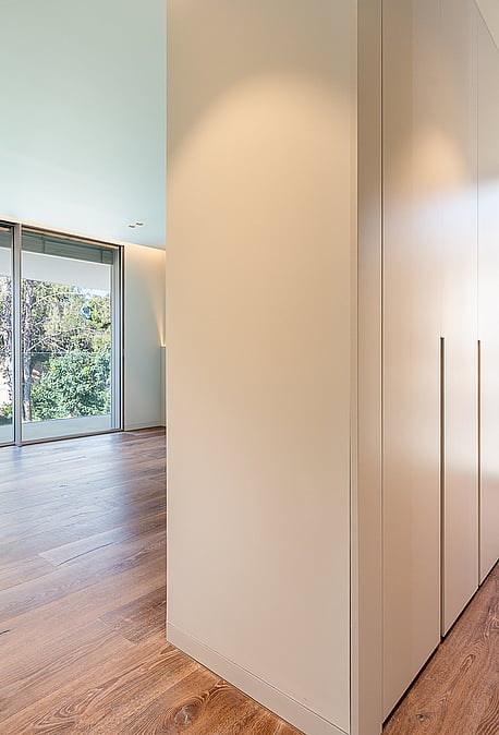 A minimalist dressing room corridor in a Santa Ponsa villa, featuring light oak wood floors, floor-to-ceiling white built-in wardrobes, and expansive glass windows overlooking greenery.