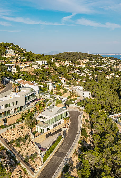 Aerial drone shot of a white contemporary hillside villa surrounded by pine trees, with a winding road and coastal horizon.
