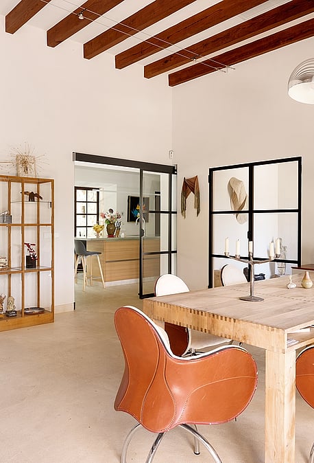 Dining area featuring a rustic wooden table, leather bucket chairs, and exposed timber ceiling beams in a spacious white room.