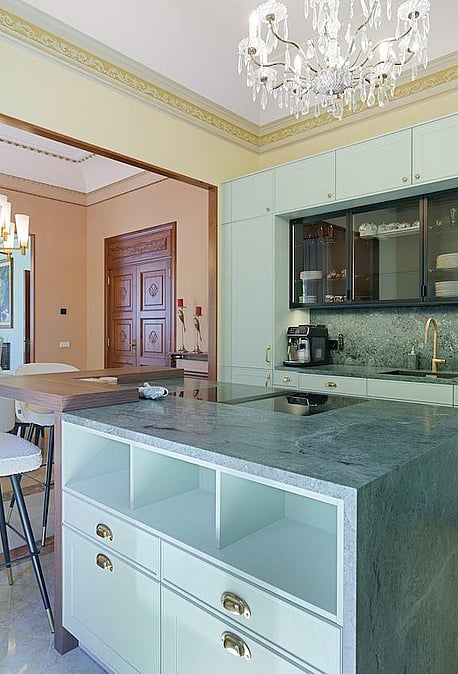 Modern kitchen with sage green cabinets, a dark grey stone island, and bar stools, leading to a dining area with a chandelier.