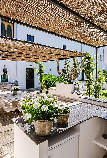 An outdoor kitchen with stone counters and a sink sits under a woven reed canopy, adjoining a paved terrace and garden.