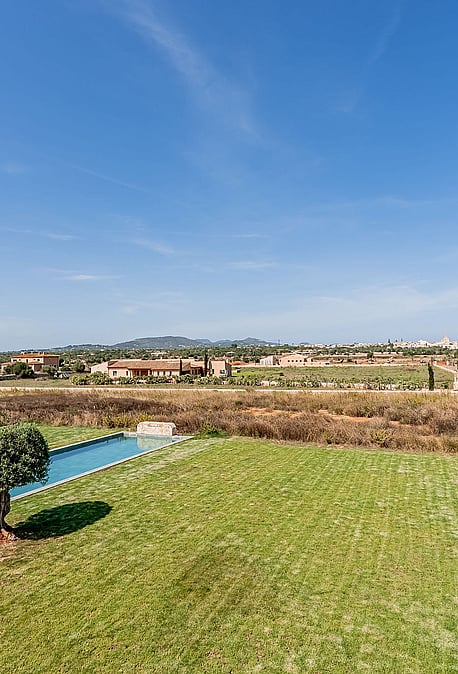An elevated view over a green lawn and rectangular pool toward distant mountains in the Santanyí countryside.