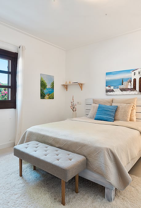 Dormitorio con cama de matrimonio, alfombra de texturas y marco de ventana de madera oscura hacia el verde.