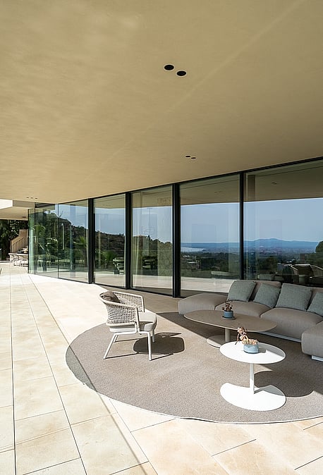 A wide, covered outdoor terrace with beige stone flooring, a modular sofa on a circular rug, and glass balustrades.