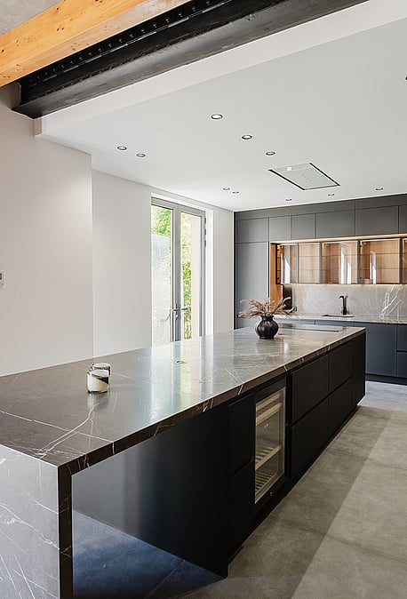 Modern open-plan kitchen in an apartment featuring a large dark stone island with waterfall edges, matte black cabinetry, and exposed wooden ceiling beams.