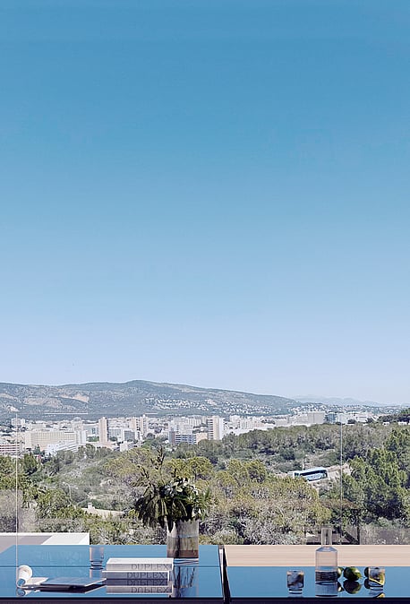 An outdoor terrace with white minimalist sofa and glass-topped table overlooking a valley, cityscape, and mountain range.