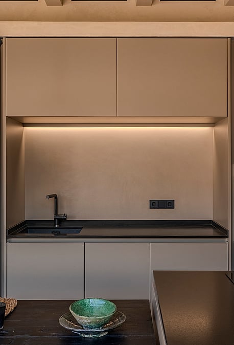 A minimalist kitchen wall featuring matte beige cabinetry, a dark countertop, integrated oven, and a sculptural textured pendant light.