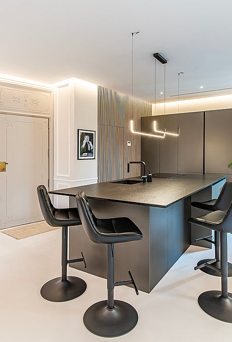 Modern kitchen featuring a dark grey central island, black swivel stools, recessed ceiling lighting, and panelled walls.