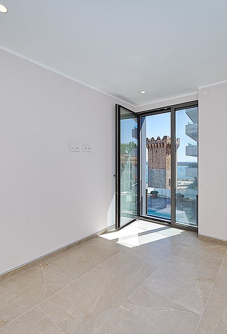 Bright unfurnished bedroom with pivot glass door opening to balcony, Torre de Paraires and harbour visible beyond