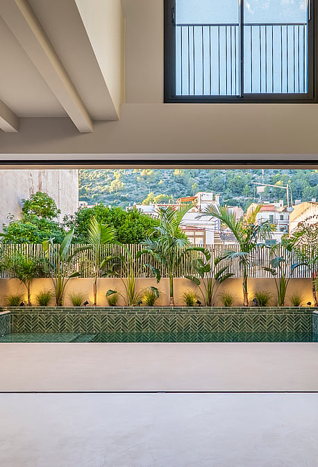 Interior view through open sliding doors to a terrace with a green tiled plunge pool and lush perimeter planting.