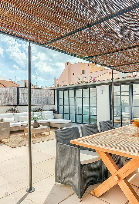 A spacious outdoor terrace with a wooden dining table, wicker chairs, and a shaded lounge area against a city backdrop.