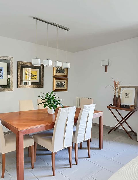 A dining room featuring a rectangular wooden table with six cream-upholstered chairs, white walls decorated with framed artwork, and a hanging pendant light.