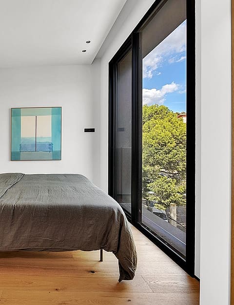 Modern bedroom with oak wood flooring, gray bedding, and large windows overlooking green trees under a blue sky.