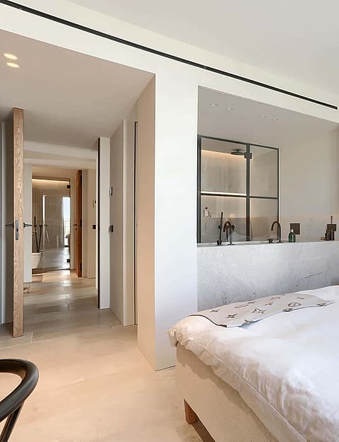Modern bedroom with a white bed, wood-paneled headboard, and an open-concept en-suite bathroom featuring marble surfaces.