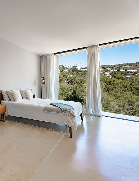 Minimalist bedroom featuring floor-to-ceiling windows, mountain views, a blue velvet chair, and polished stone floors.