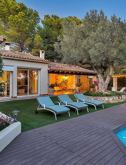 Outdoor swimming pool with blue water and timber decking, surrounded by garden, olive tree, and shaded stone terrace.