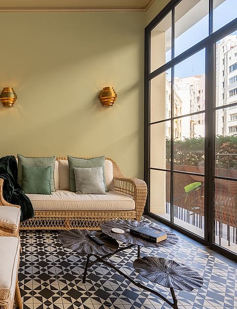 Interior sunroom with woven rattan furniture, a large palm leaf mural, and black-framed glass windows overlooking the city.
