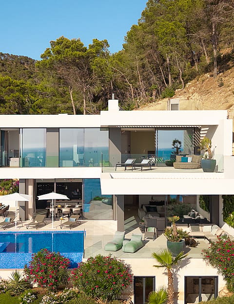 Aerial view of a white, multi-level modern villa built into a hillside, featuring a blue tiled infinity pool and sun loungers.