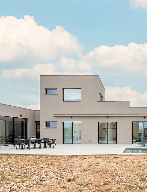 Rear view of a modern two-story beige villa with a swimming pool, paved terrace, and a separate covered outdoor lounge area.
