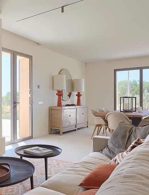An open-plan living and dining room with a built-in fireplace, light-toned sofas, two round coffee tables, and a wooden dining table in the background, facing exterior mountain views.