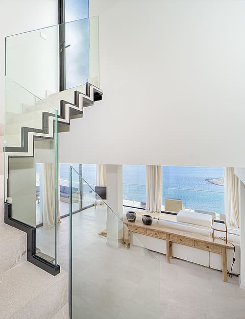 A white stone staircase with a clear glass railing connects to an upper floor, overlooking an open-plan living area with sea views.