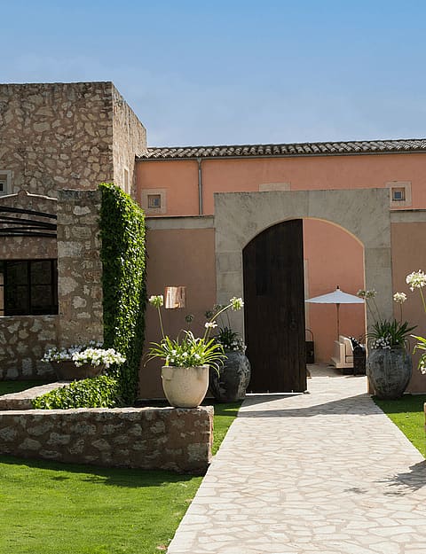 A stone paved path leads through an arched wooden gate into a courtyard of a traditional Finca in Algaida.