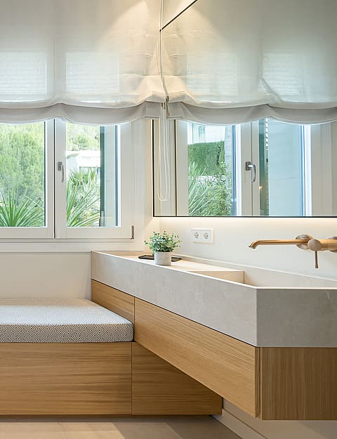 Contemporary bathroom with a white built-in tub, stone-textured walls, oak wooden vanity, and a window with white Roman blinds.