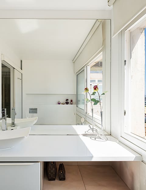 A modern white bathroom vanity with a square vessel sink and a large wall mirror overlooking a sunlit coastal city scene.