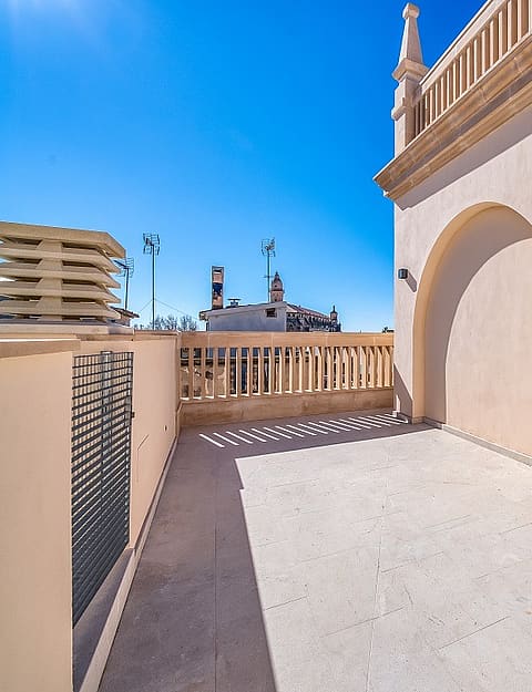 A rooftop terrace featuring light-colored stone flooring, stucco walls with a decorative arch, and stone balustrade railings under a clear blue sky.