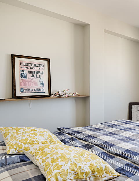 A bedroom featuring a bed with a blue plaid quilt, a floating wooden shelf with framed artwork, and a window overlooking city buildings.