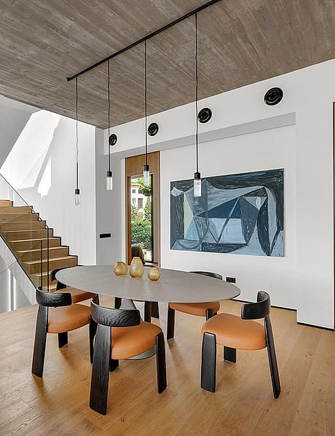 Dining area with oval table and wooden chairs, adjacent to a wooden staircase under a raw concrete ceiling.