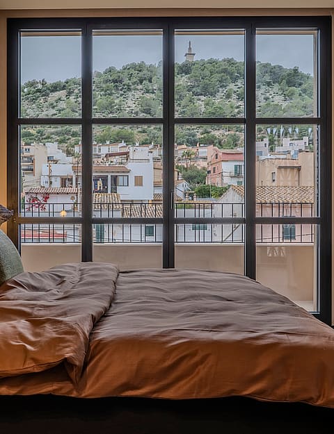A contemporary bedroom featuring a dark bed frame with a textured wooden headboard, terracotta bedding, and a large metal-framed window looking out onto a hillside.