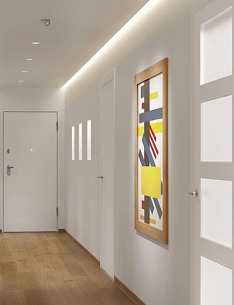A wide interior hallway with white walls, light oak flooring, and recessed ceiling lighting leading to an entrance door.
